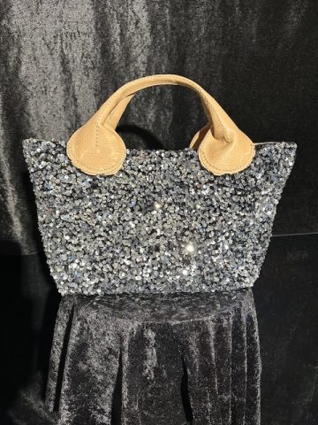 Silberne Paillettentasche mit beigefarbenen Griffen auf dunklem Stoffhintergrund.
